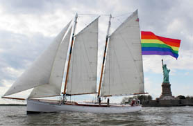 Schooner Adirondack sailing with a 400 square foot PRIDE Flag in front of the Statue of Liberty on a cloudy day