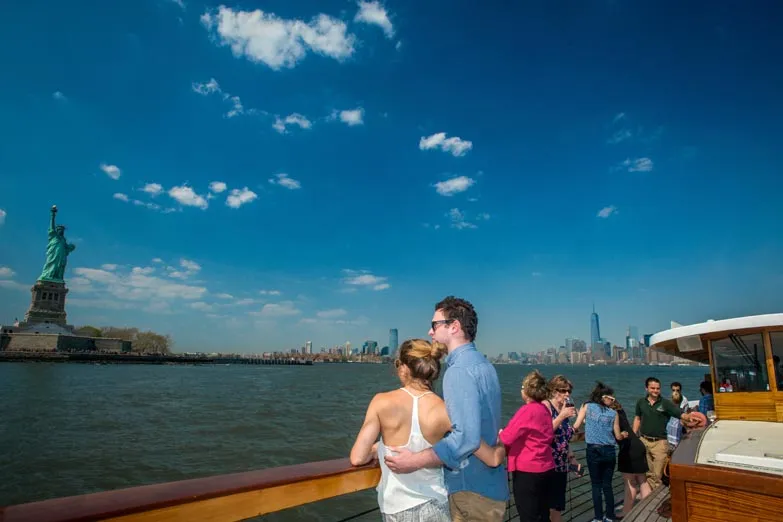 Couple on the bow of a boat in summer attire with the Statue of Liberty and NYC Skyline in the background
