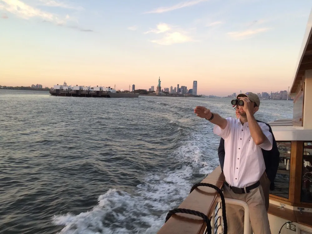 A man holding binoculars on a boat and pointing out over the water with the Statue of Liberty behind them