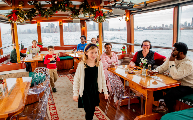 Interior salon of a classic yacht with Holiday decor and customers enjoying food and beverages with the NYC Skyline in the background.