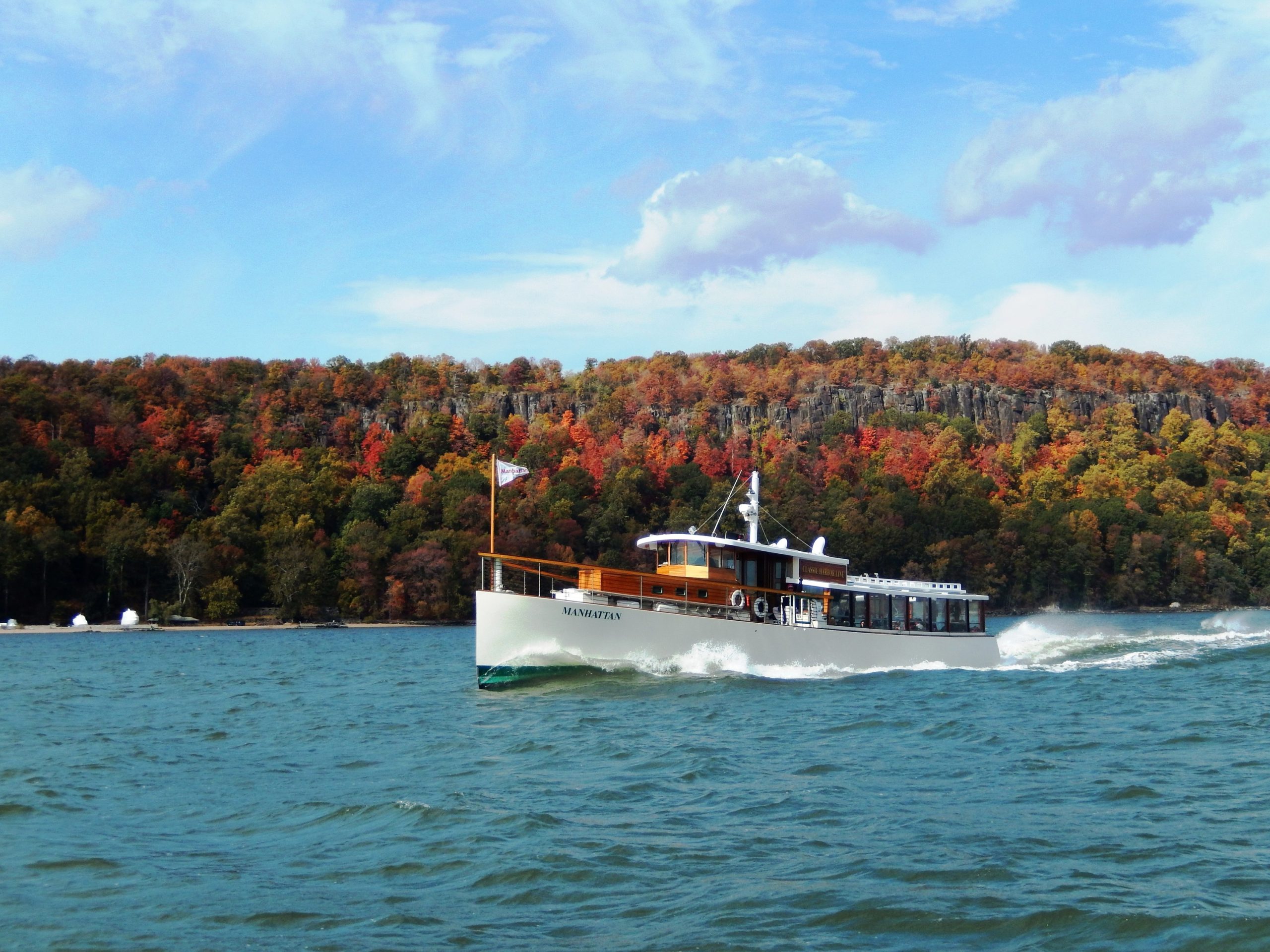 Motor yacht Manhattan II cruises on the Hudson past the vibrant fall foliage on the palisades in the fall.