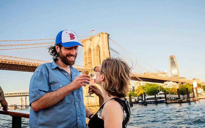 ChampCheeseWeb2 Couple standing on the deck of a classic yacht, enjoying drinks with the Brooklyn Bridge in the background.