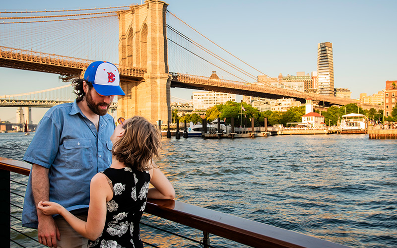 Couple on the bow of a boat with the Brooklyn Bridge in the background.