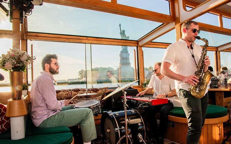 Three person jazz band playing on the inside of a classic yacht with the Statue of Liberty in the background.