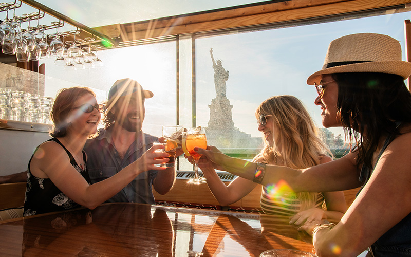 Group of friends clinking cocktails seated at a table on a yacht with the Statue of Liberty in the background.