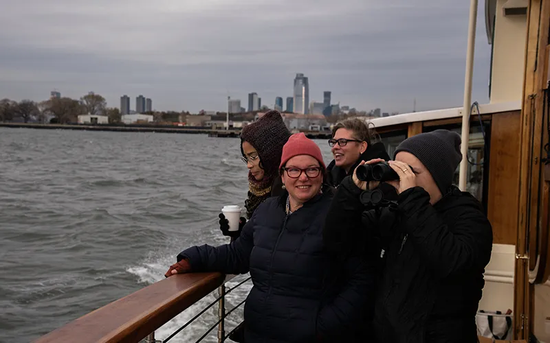 four people looking out off the bow of a boat with NYC in the background.