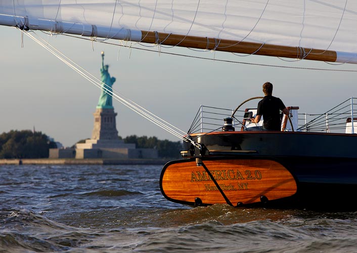 Stern shot of schooner America 2.0 with Captain driving the yacht past the Statue of Liberty