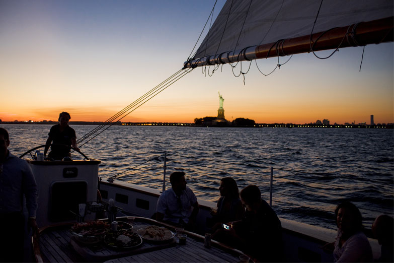 Captain and guests in the cockpit of Schooner America 2.0 at sunset with the Statue of Liberty in the distance