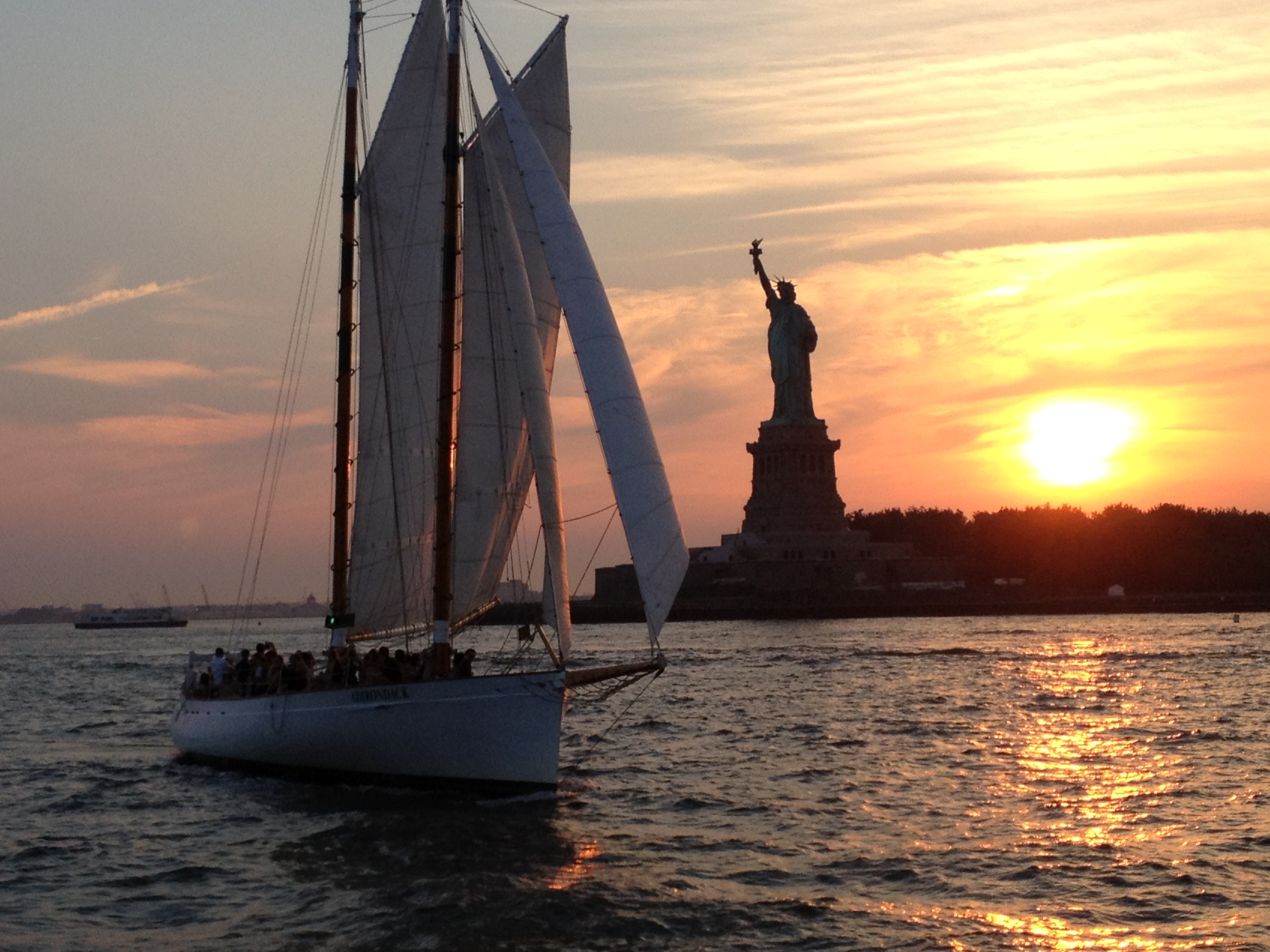 Full boat photo of the Schooner Adirondack sailing past the Statue of Liberty as the sun dips beyond the horizon