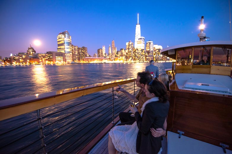 A couple sitting on the wrap around bench on the bow of the yacht Manhattan for a city lights cruise with the city lit up at night 