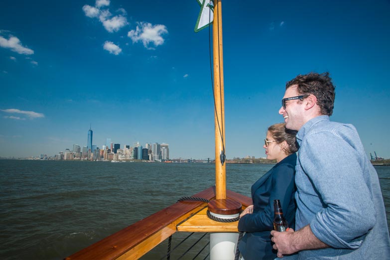 Couple looking off the bow of Yacht Manhattan with the Manhattan Flag above their heads and the skyline in the background
