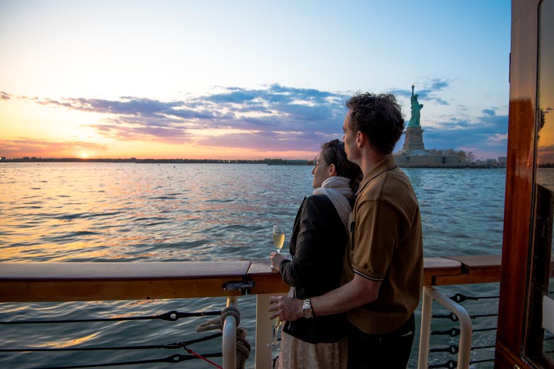 A couple standing, holding each other as they look off to the Statue of Liberty for a sunset cruise