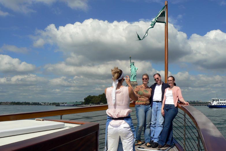 Crew member taking a photo of a family in front of the Statue of Liberty on the bow of Yacht Manhattan on a sunny afternoon day